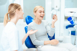 A woman sits in a dental chair, conversing with another woman, likely a dental professional, in a clinical setting.